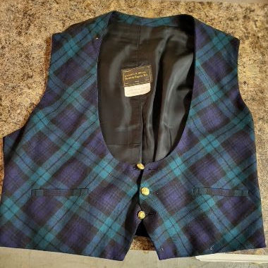 A men's waistcoat vest with a blue and purple tartan plaid pattern, laid flat on a speckled granite countertop. The vest features a black satin inner lining, three gold-colored buttons, and two front welt pockets.